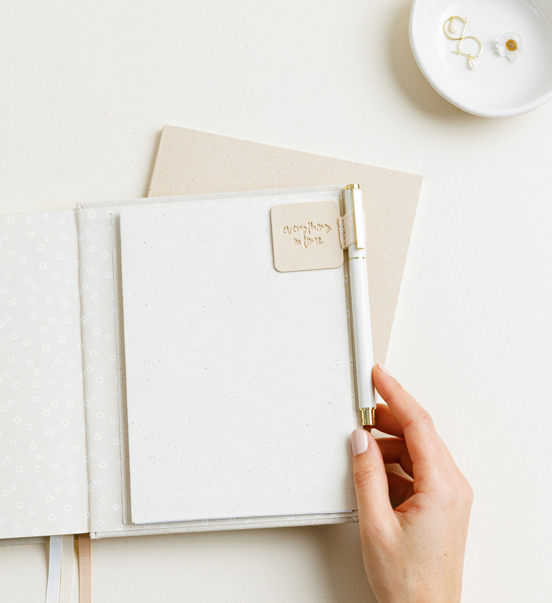 Minimal flatlay of a soft-toned open journal with floral lining, speckled paper, and a cream pen held by a hand, alongside a ceramic dish with gold earrings and a daisy detail